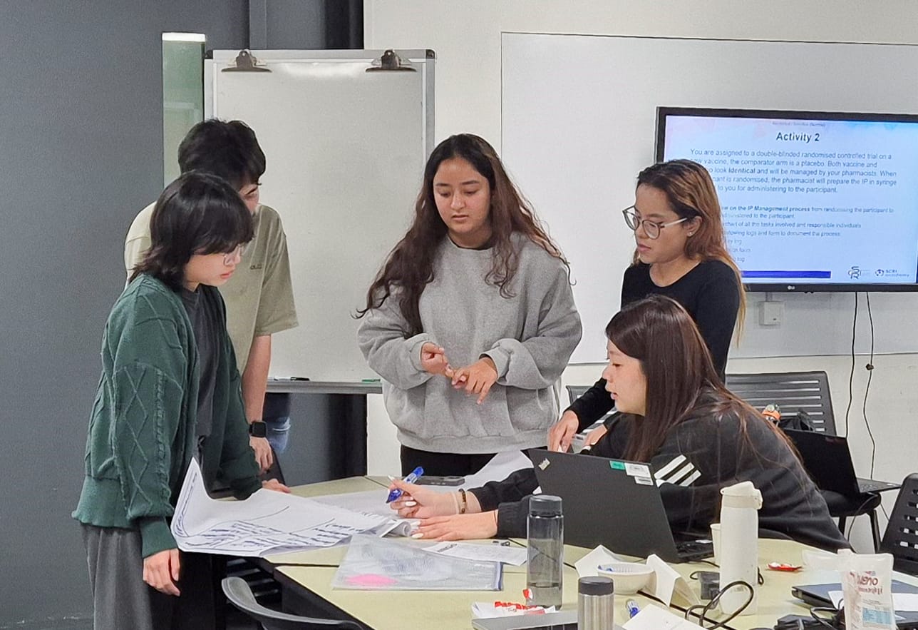 Five people gathered around a table with papers and laptops in a classroom, discussing a task.